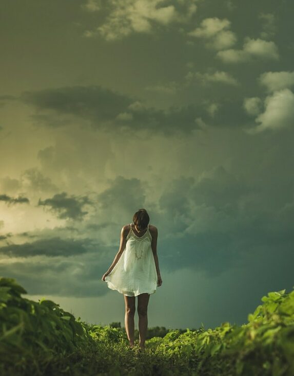 woman wearing white dress standing on hill with green grass under white cloudy sky